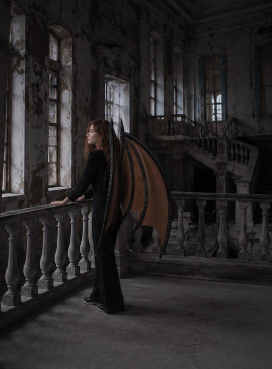 Person wearing a dragon wings costume standing in an old, abandoned building with large windows.