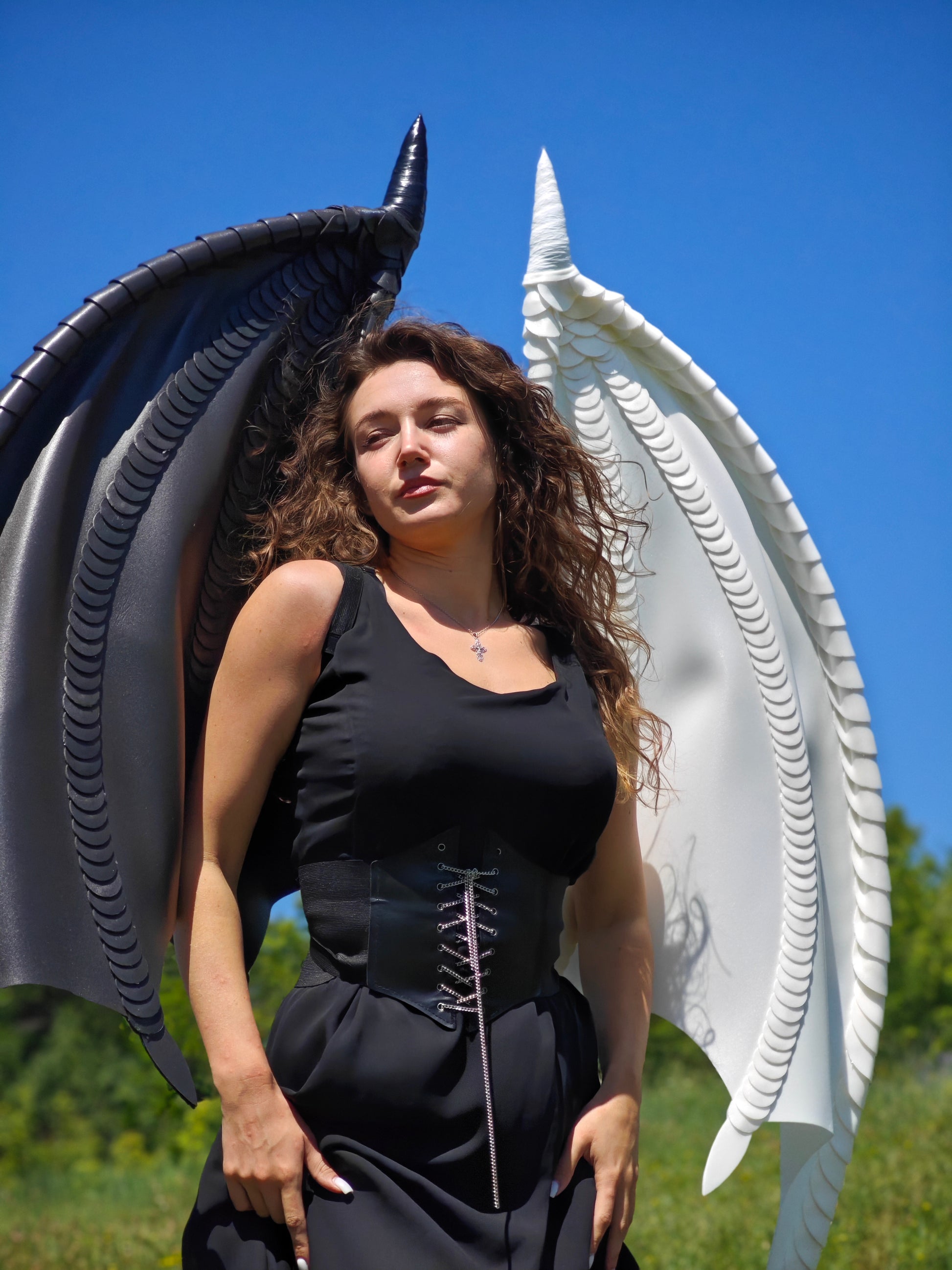 Woman in a black and white dragon wings standing between large, wing-like structures against a blue sky.