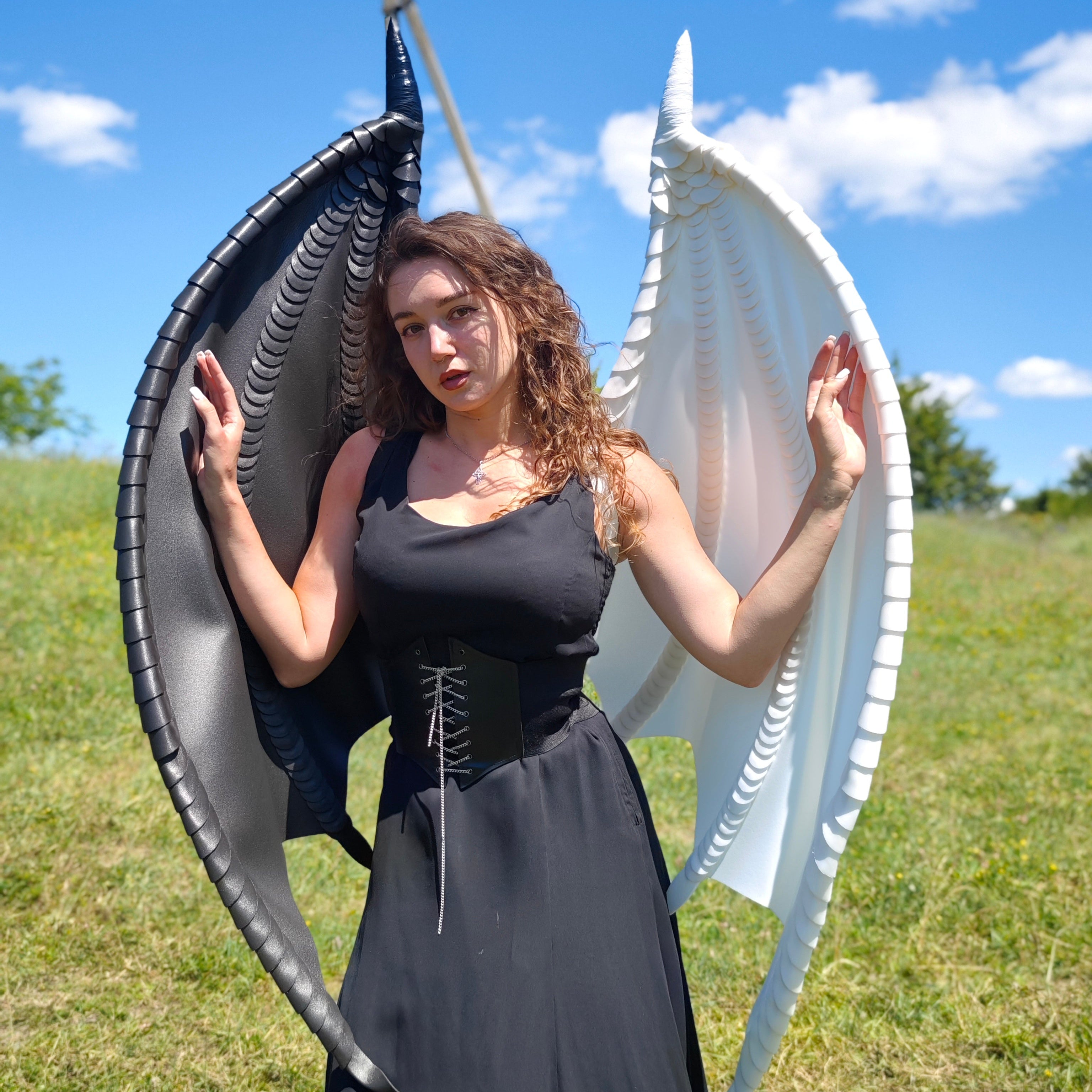 Woman in a black dress with large, decorative wings standing outdoors on a grassy field.