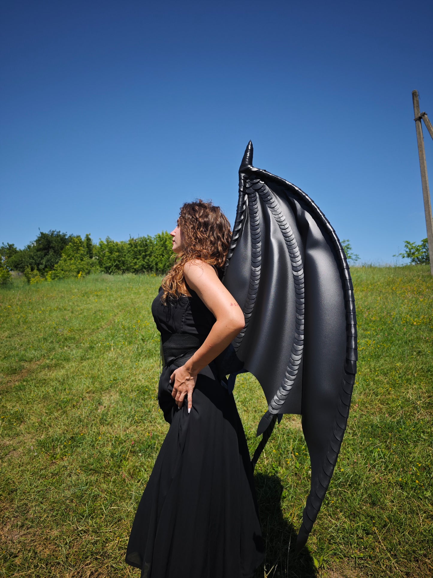 Person wearing a large black inflatable dragon wing in an open field with a clear blue sky.