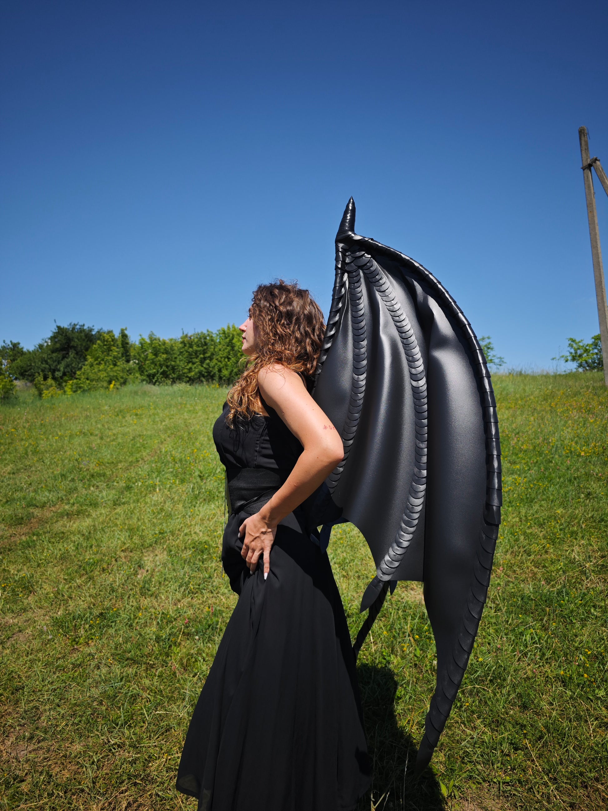 Person wearing a large black inflatable dragon wing in an open field with a clear blue sky.