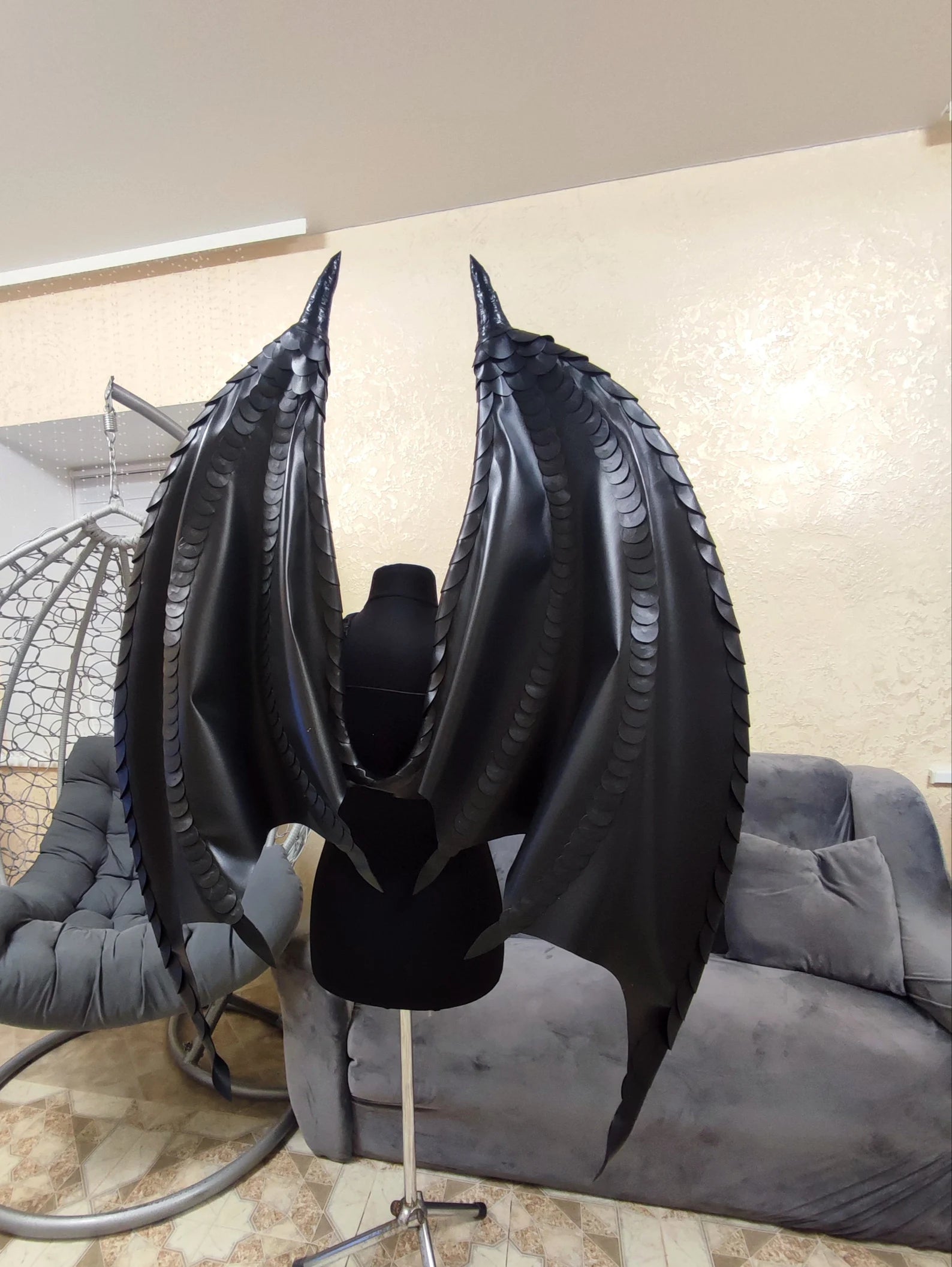Black dragon wings costume on a stand in a room with a gray chair and white wall.