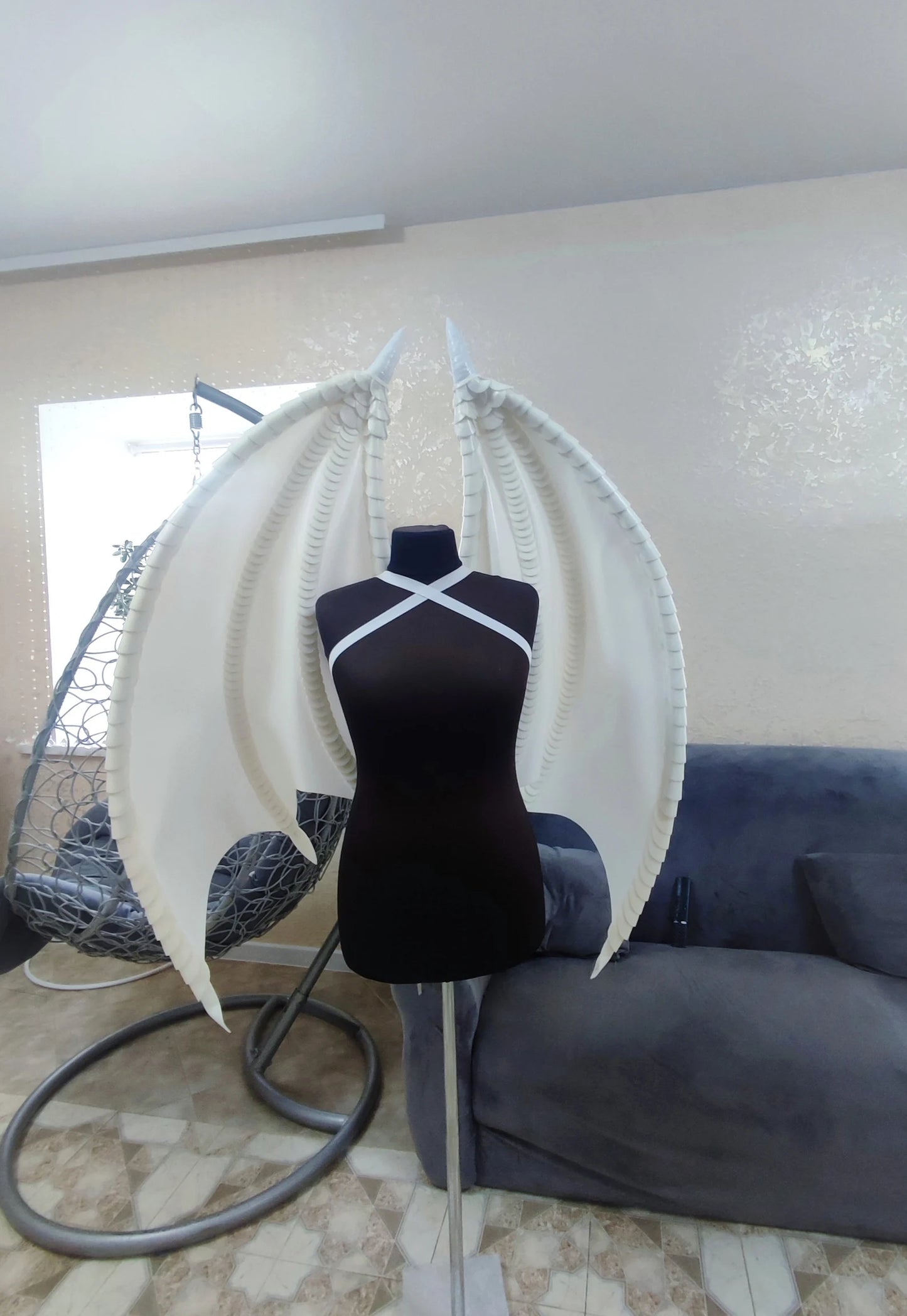 Mannequin with large white wings on a stand in a room with a couch and window.