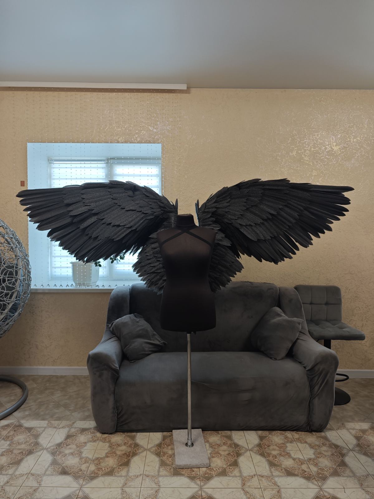 Mannequin with large black wings on a gray sofa in a room with beige walls and a window.