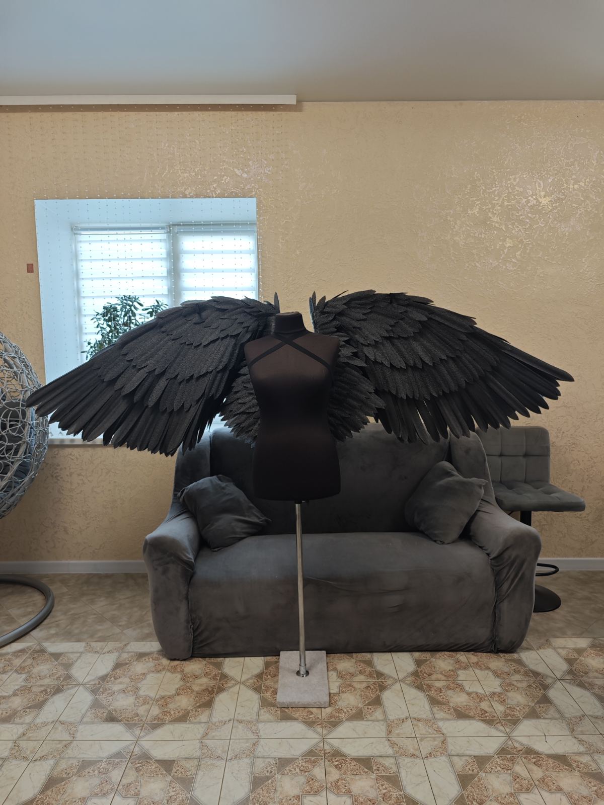 Mannequin with large black wings on a gray sofa in a room with beige walls and a window.
