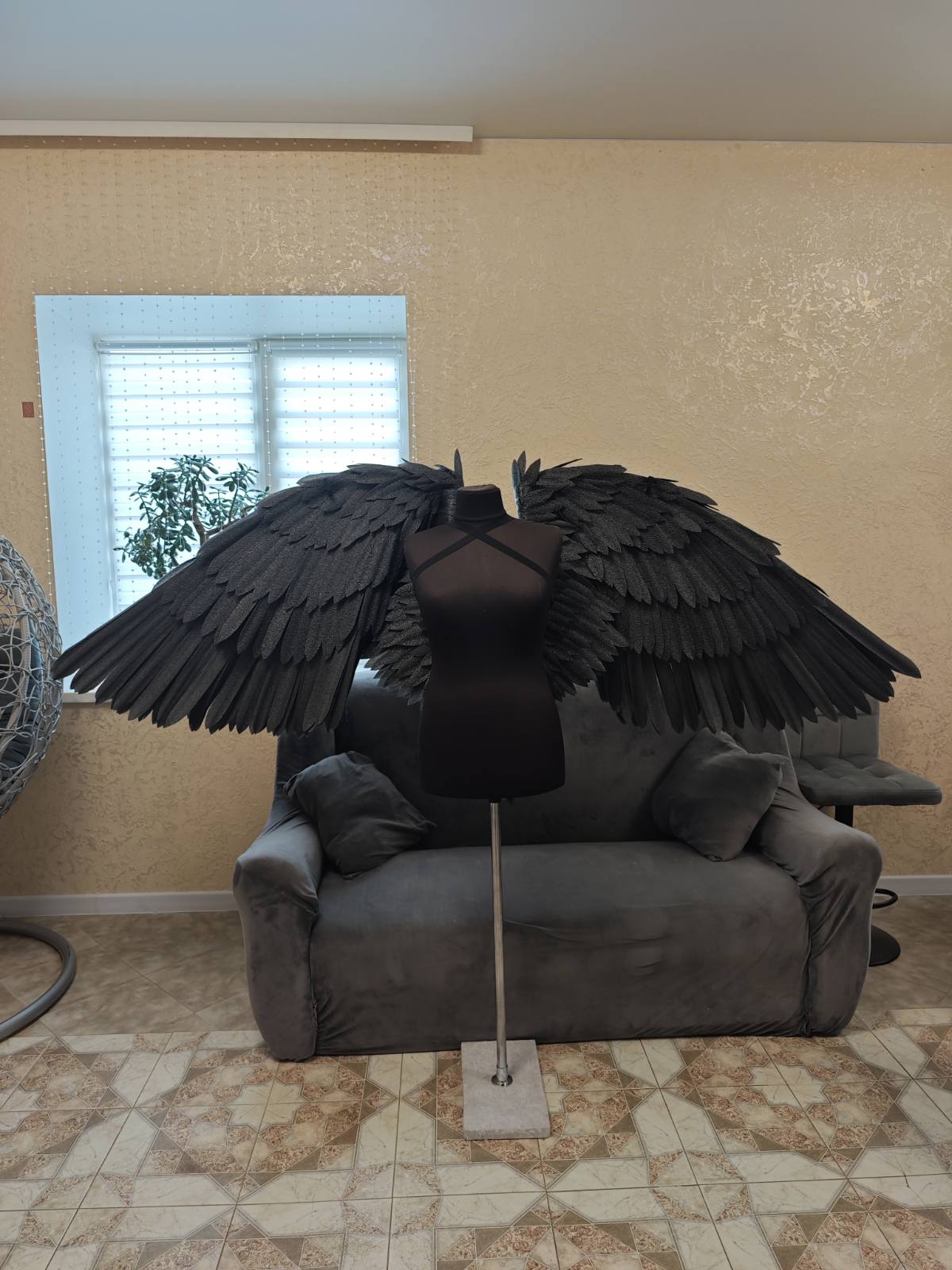 Mannequin with large black wings on a gray couch in a room with tiled floor and window.