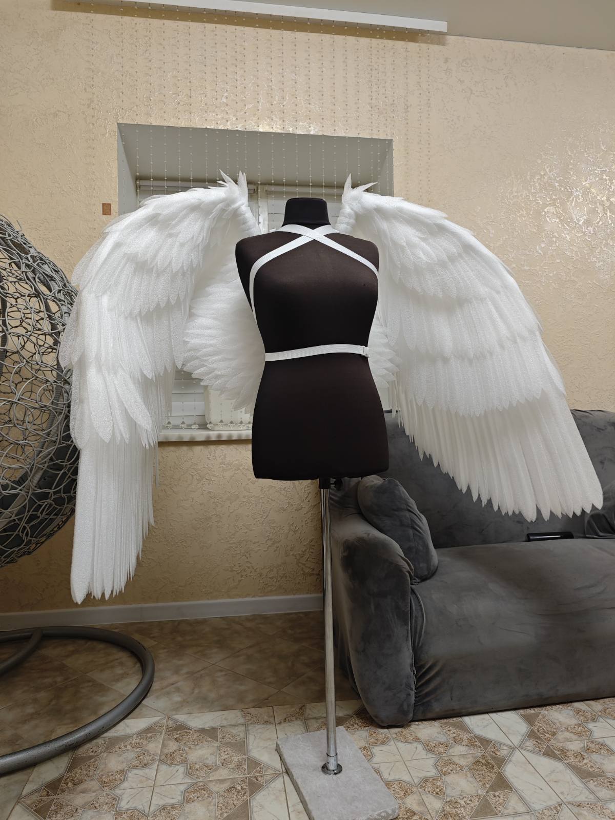 Mannequin with white angel wings in a room with a textured wall and tiled floor.