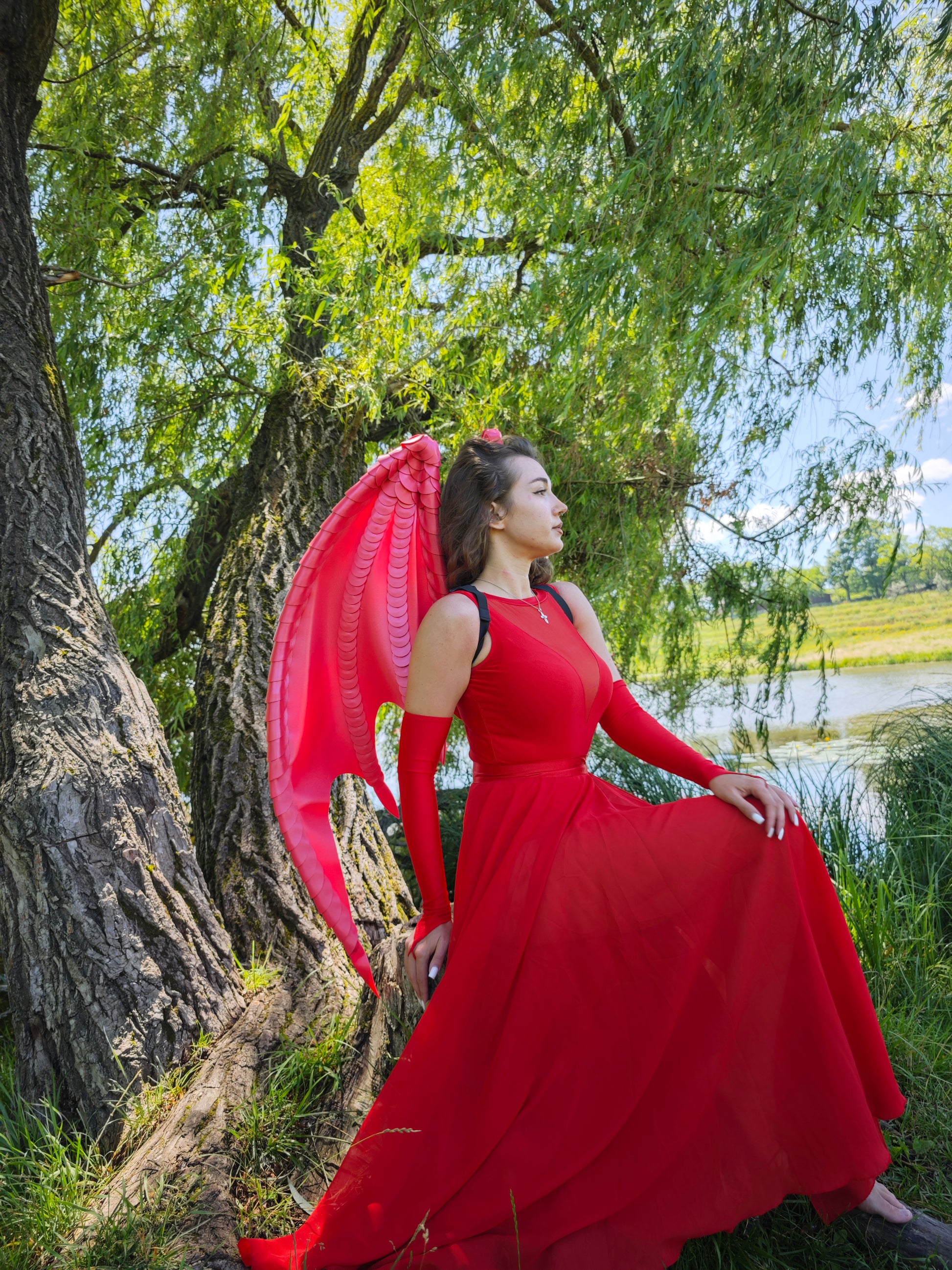 Woman in a red dress with a red dragon wings standing by a tree near a body of water.