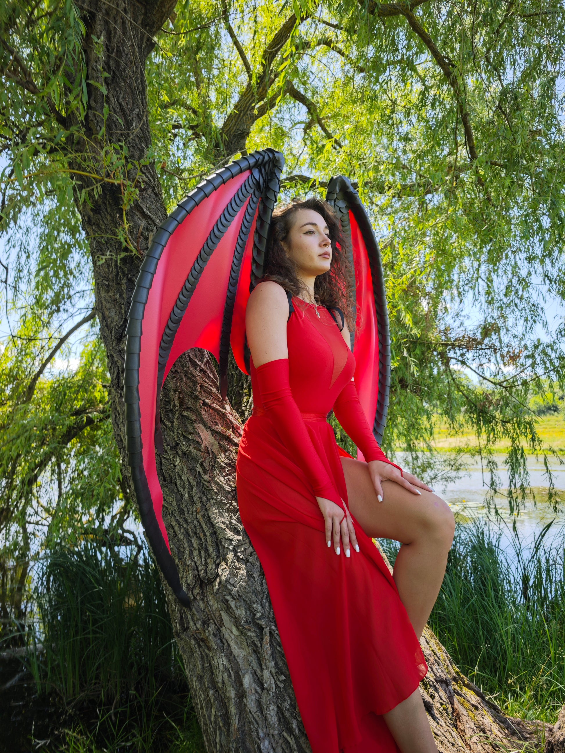 Person in a red dress with large red black wings standing against a tree by a body of water.