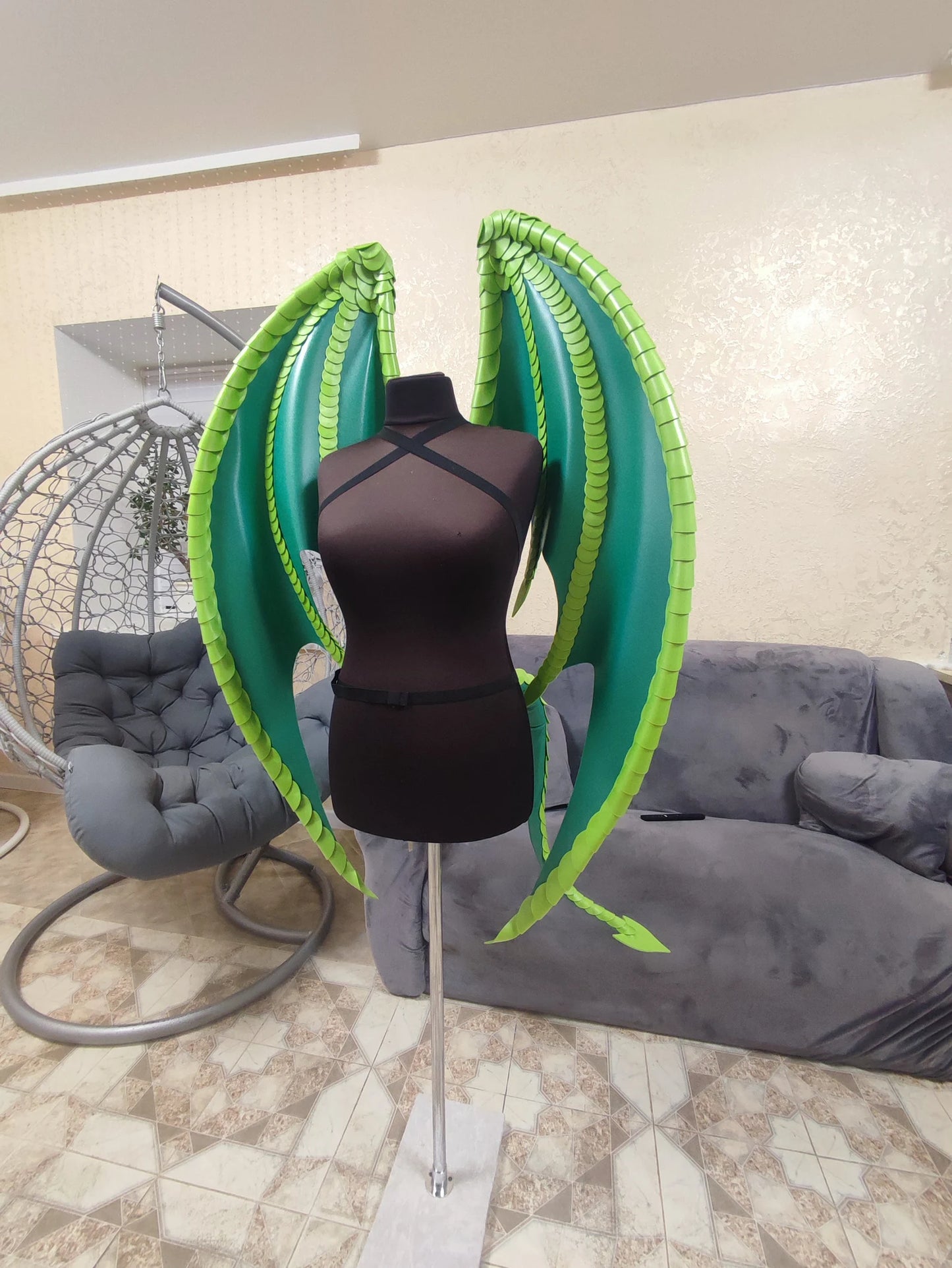 Mannequin with green wings in a room with a gray couch and chair.