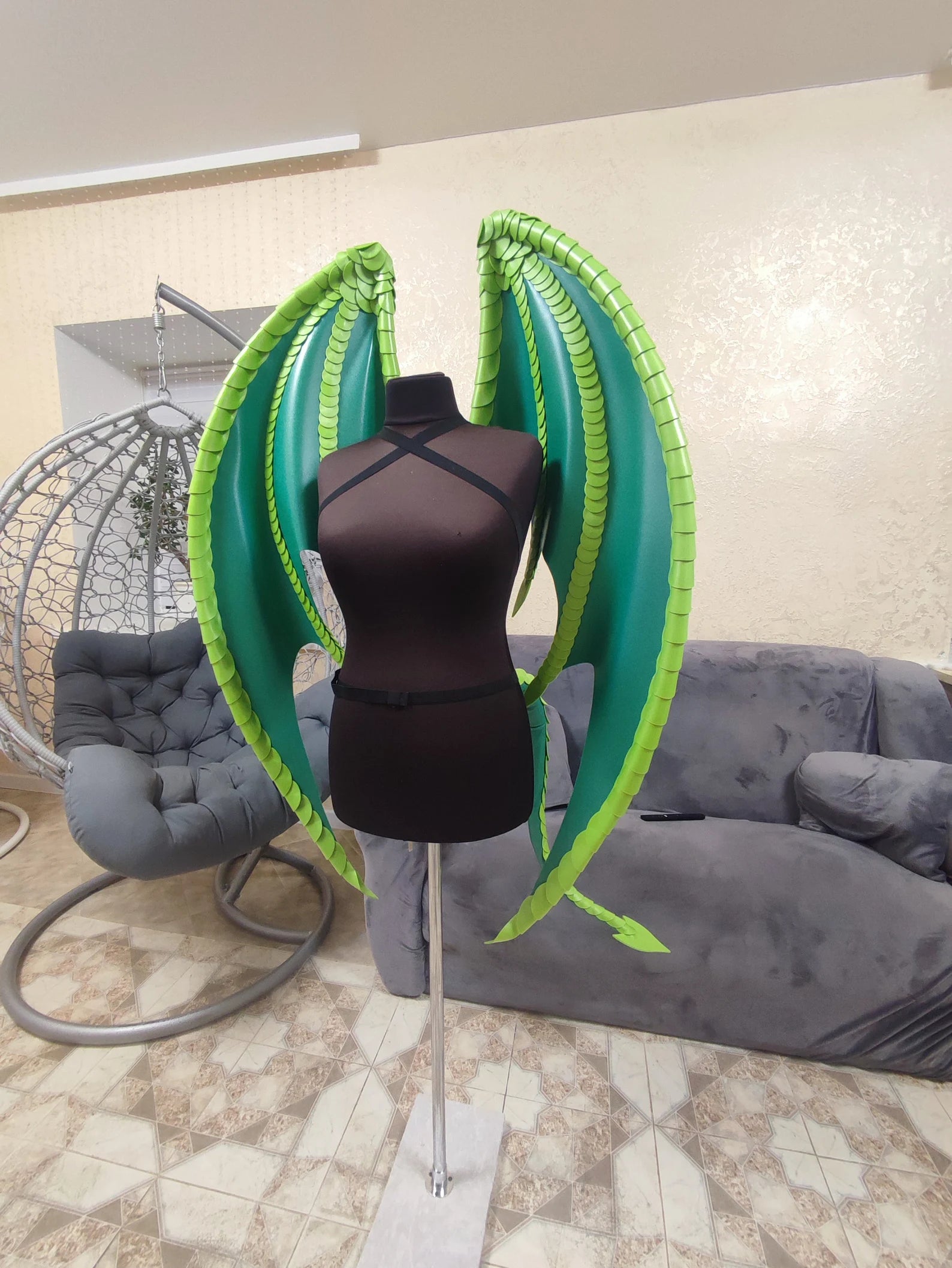 Mannequin with green wings in a room with a gray couch and chair.