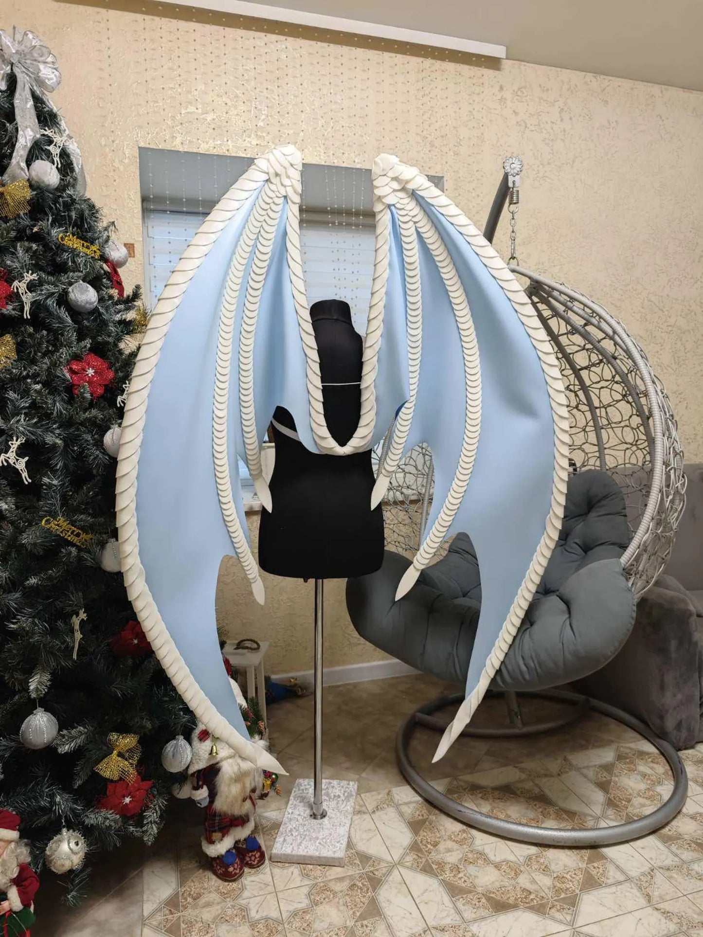 Large blue and white dragon halloween wings on a mannequin in a room with a Christmas tree and chair.