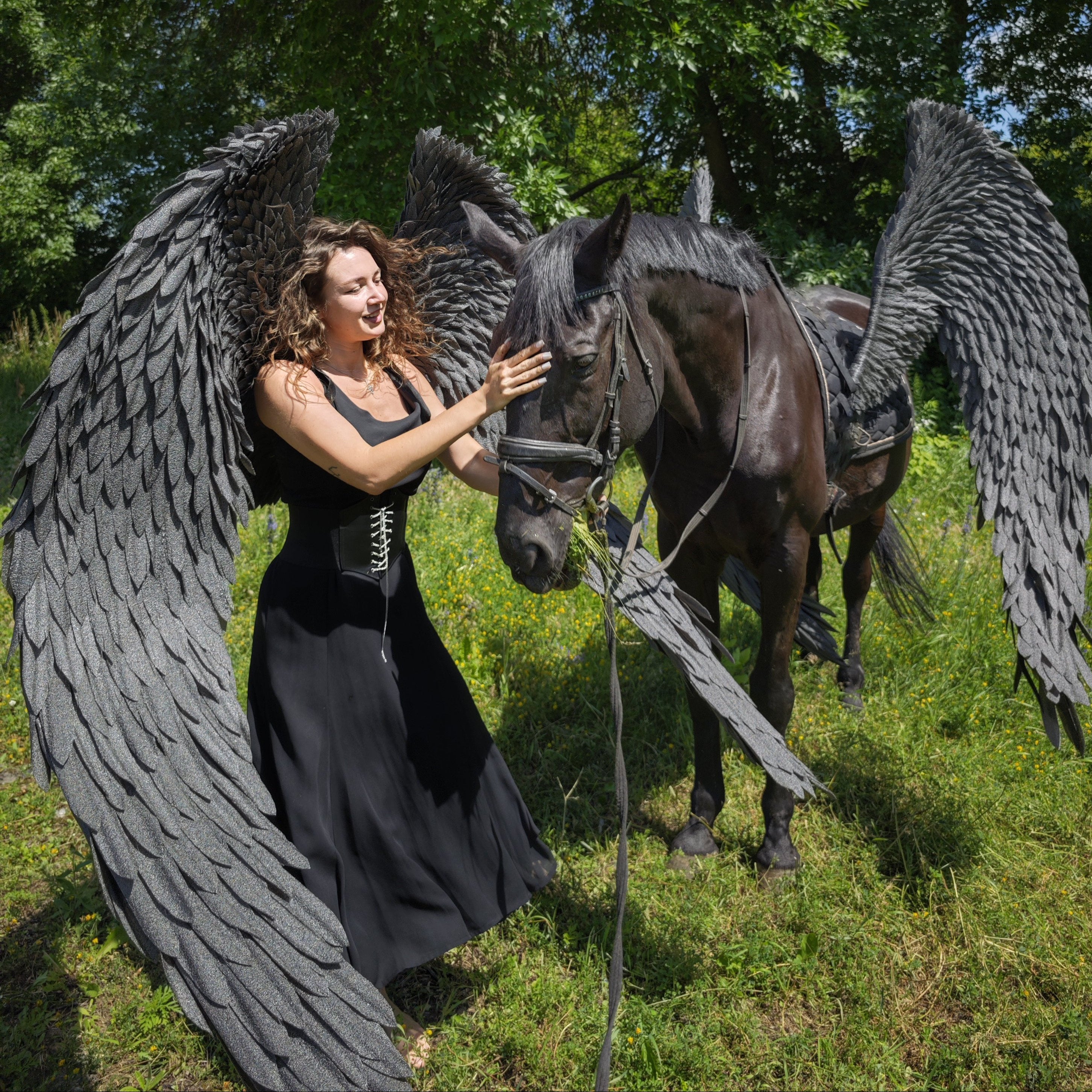 Woman with large black angel wings standing next to a horse with similar wings in a forest setting