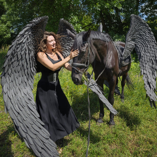 Woman with large black angel wings standing next to a horse with similar wings in a forest setting