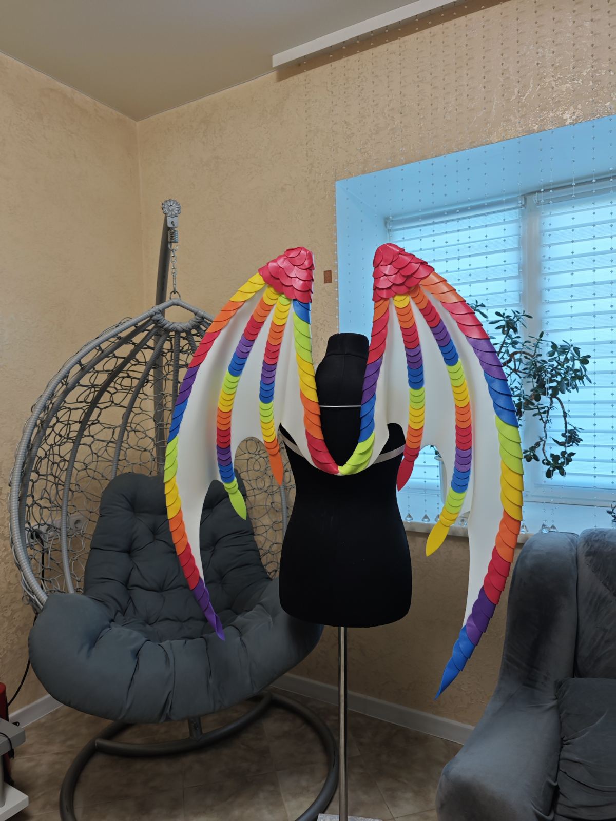 Colorful butterfly wings on a mannequin stand in a room with a chair and window.