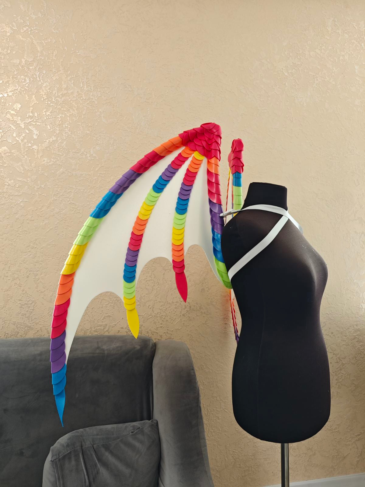 Mannequin with colorful rainbow wings extensions against a beige wall.