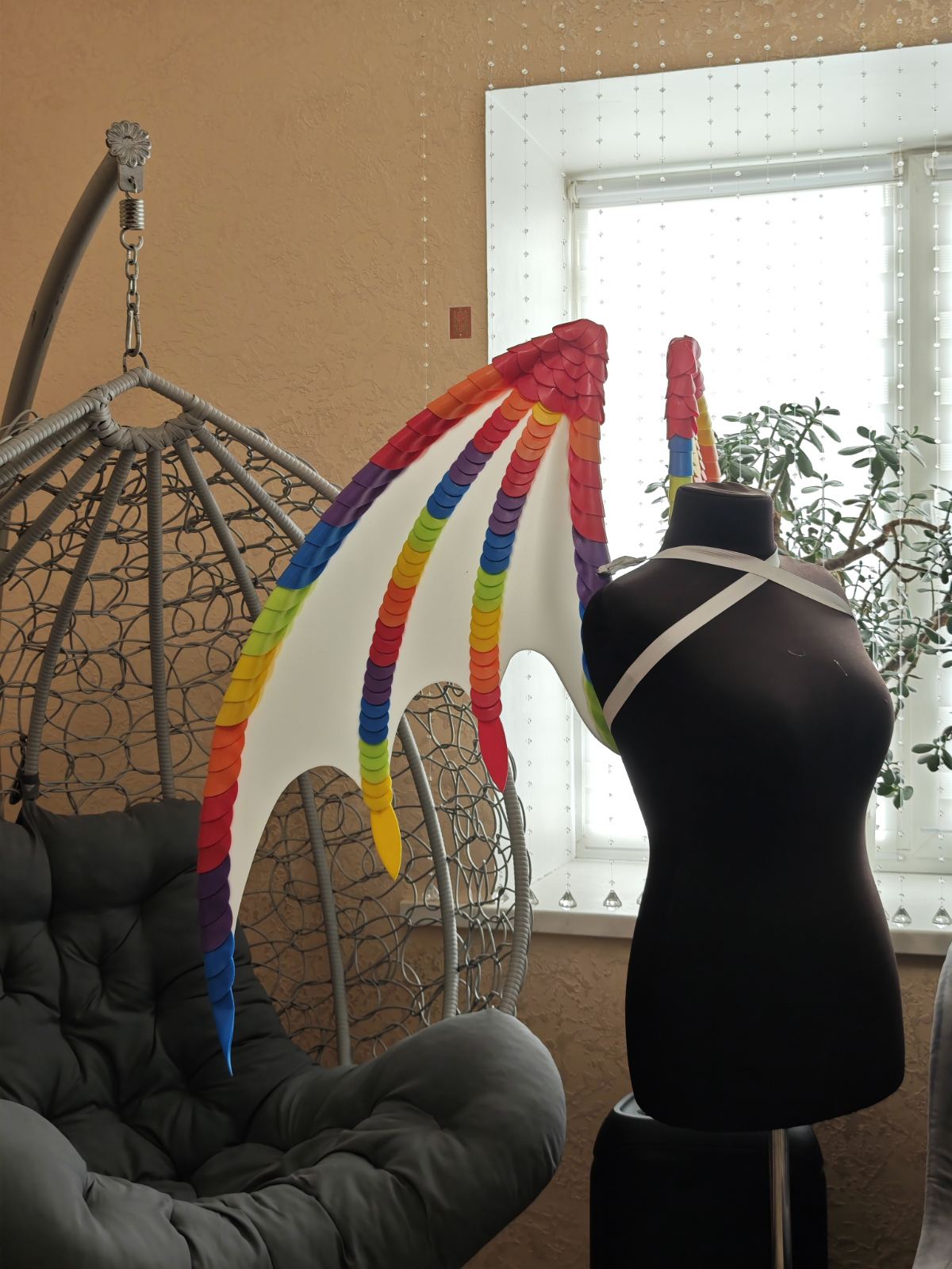 Mannequin wearing a colorful rainbow wings in a room with a hanging chair and window.