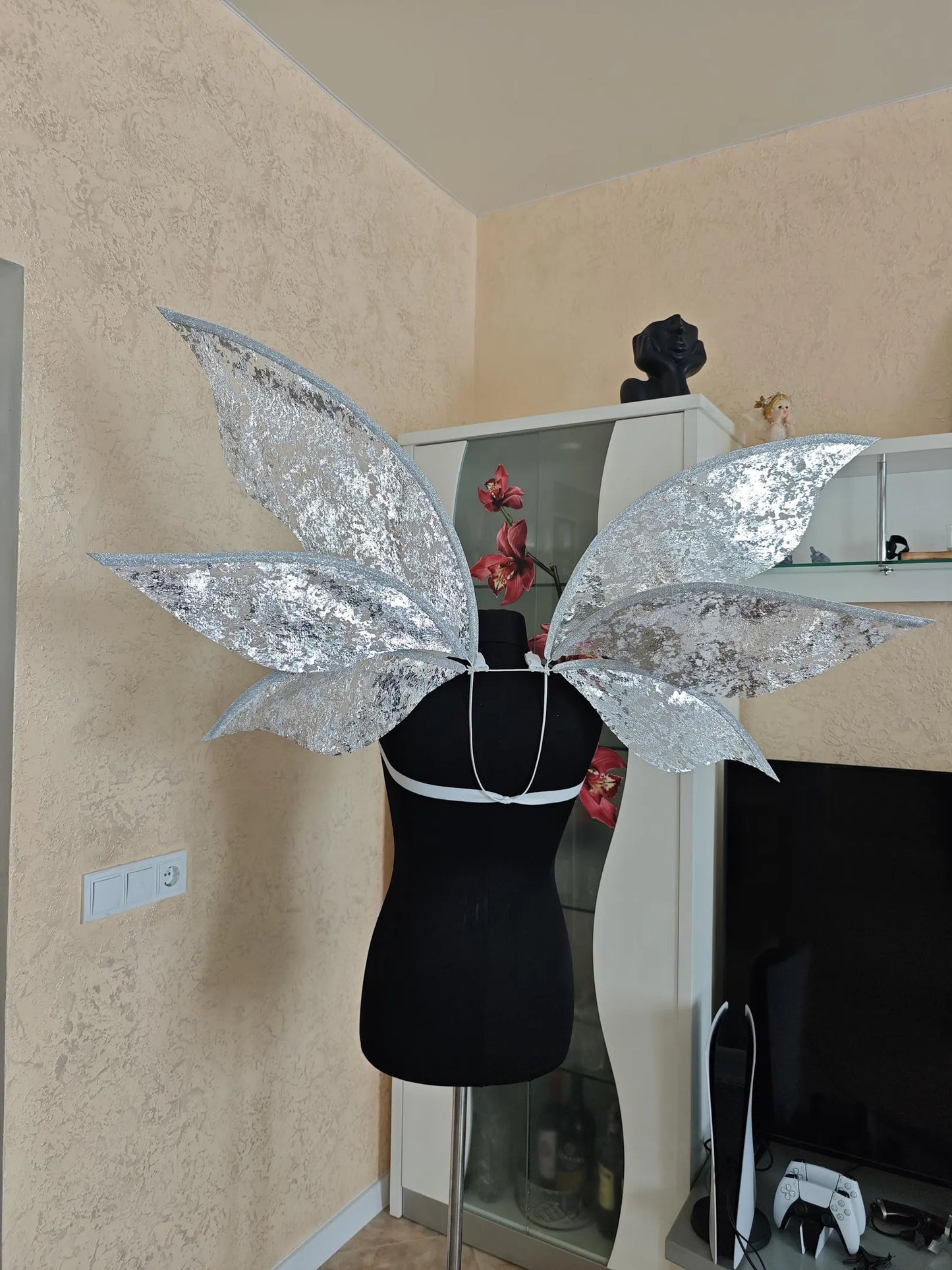 Silver butterfly wings on a mannequin against a neutral wall.