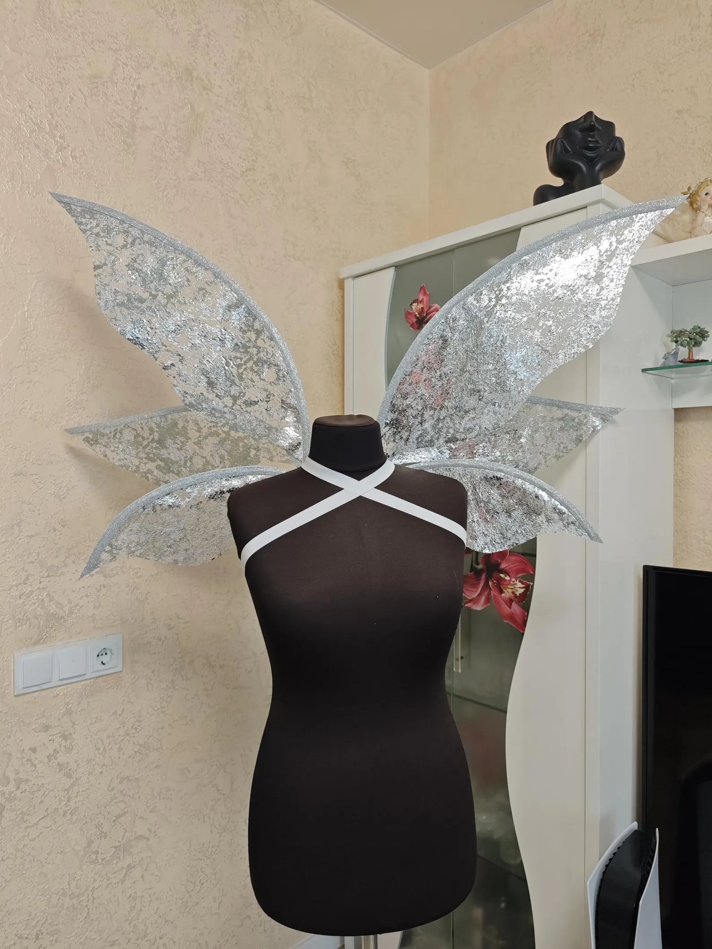 Silver butterfly wings on a mannequin against a neutral wall.