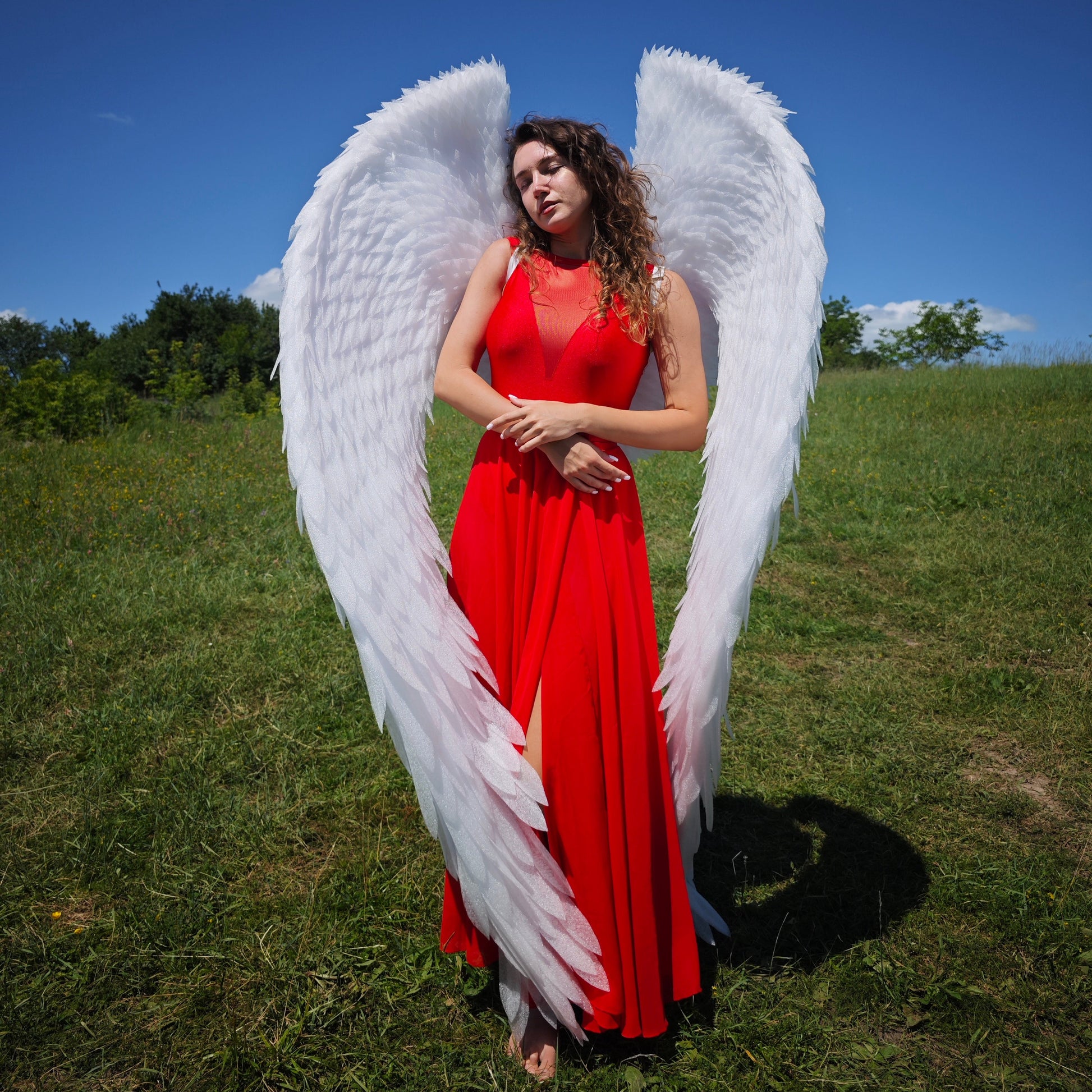 Woman in a red dress with large white wings standing on a grassy field with a blue sky.