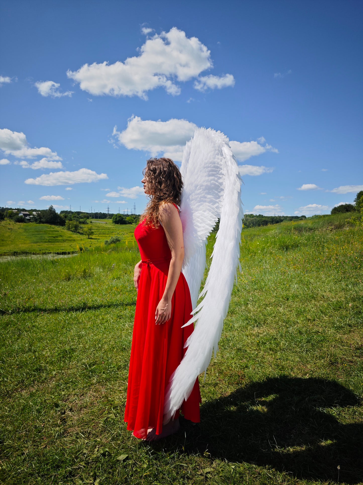 Person in a red dress with large white wings standing in a grassy field under a blue sky.