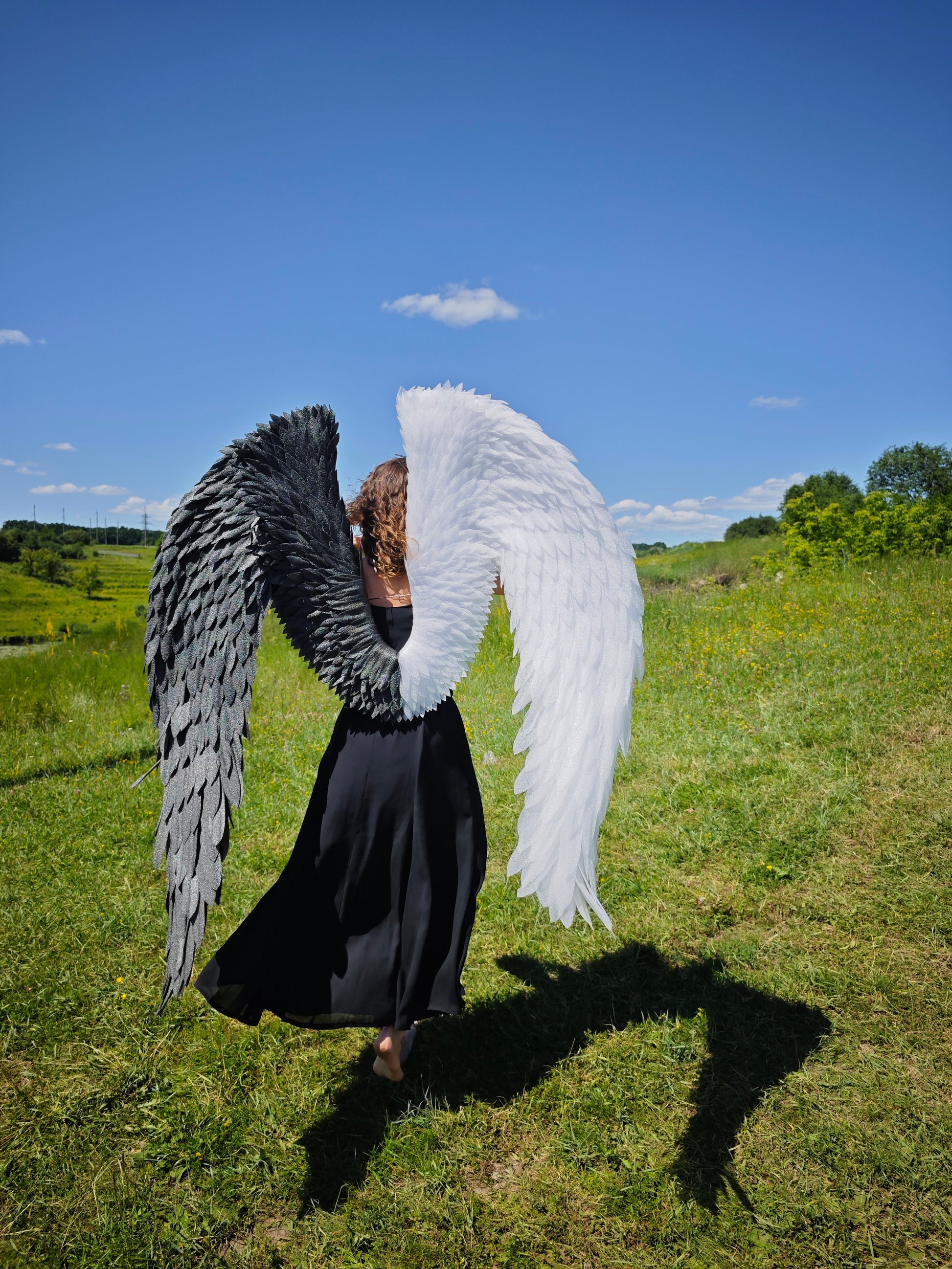 Person wearing large angel wings standing in a grassy field with a blue sky.