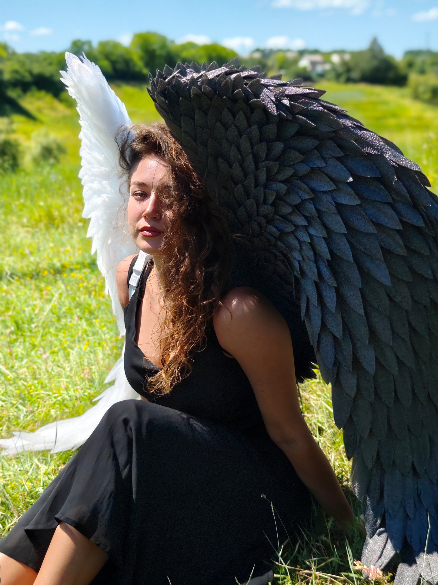 Person with large black and white angel wings sitting in a grassy field.