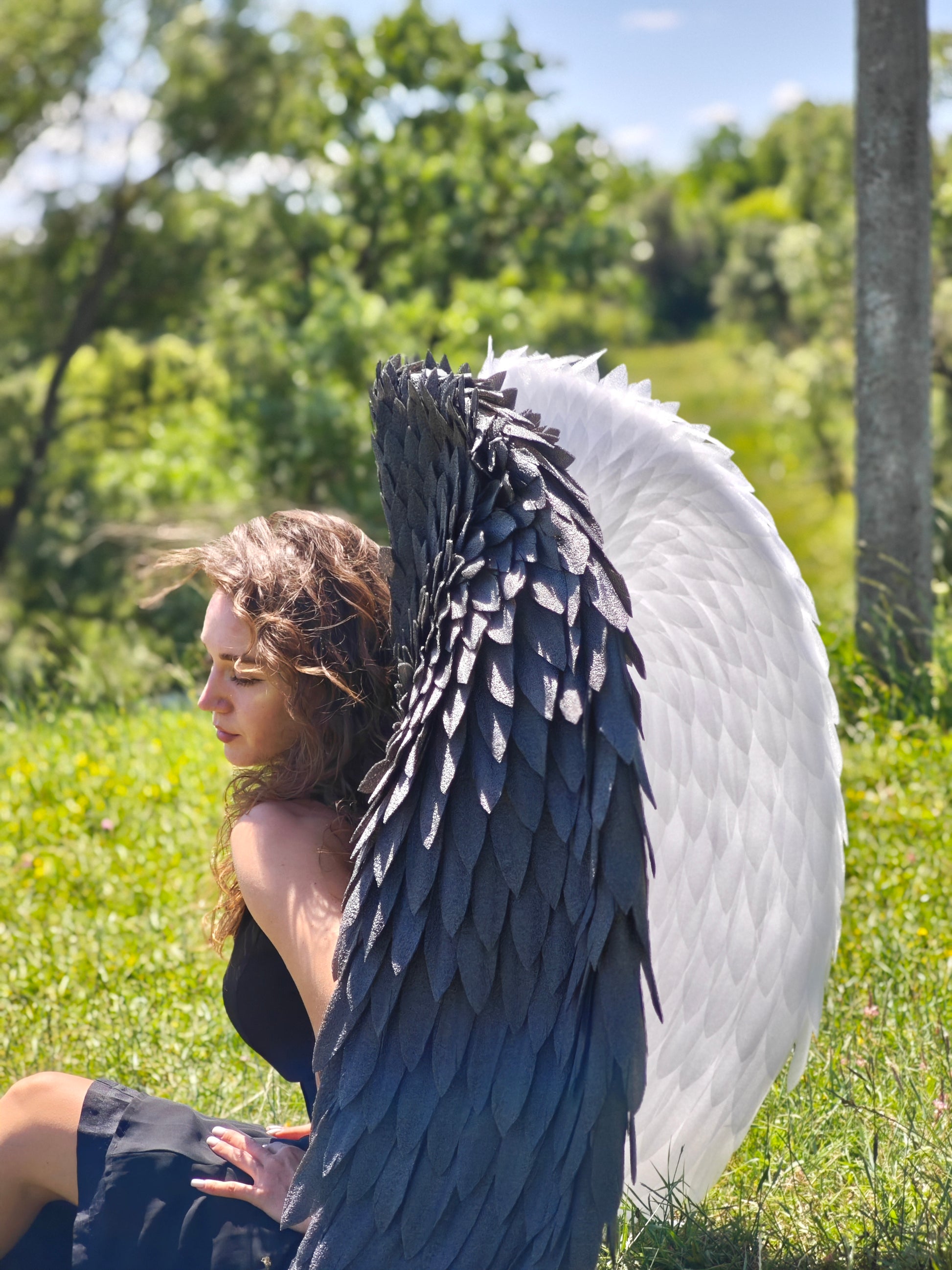 Person with large black and white wings sitting in a grassy field with trees in the background
