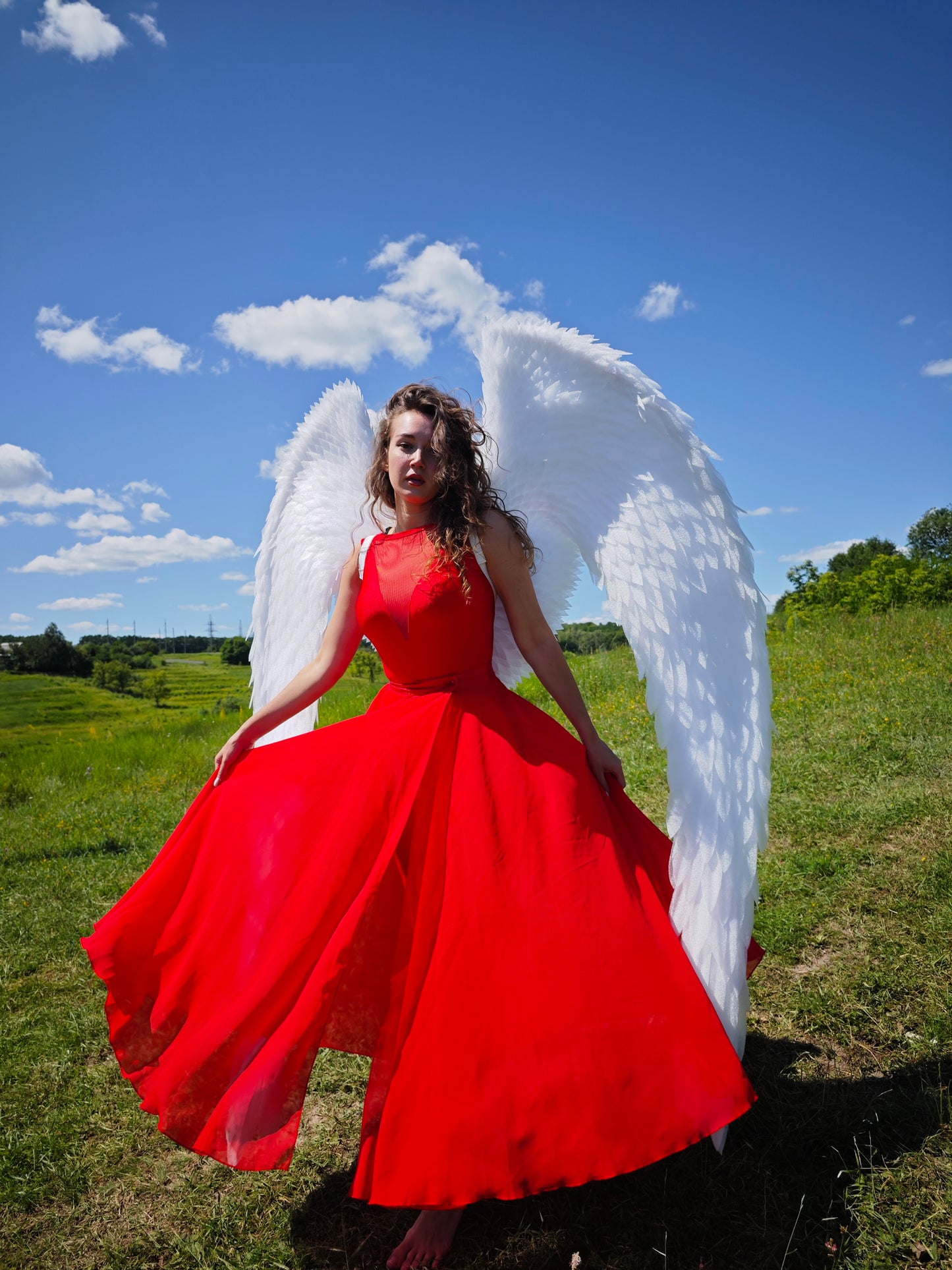 Person in a red dress with large white wings standing in a grassy field under a blue sky.