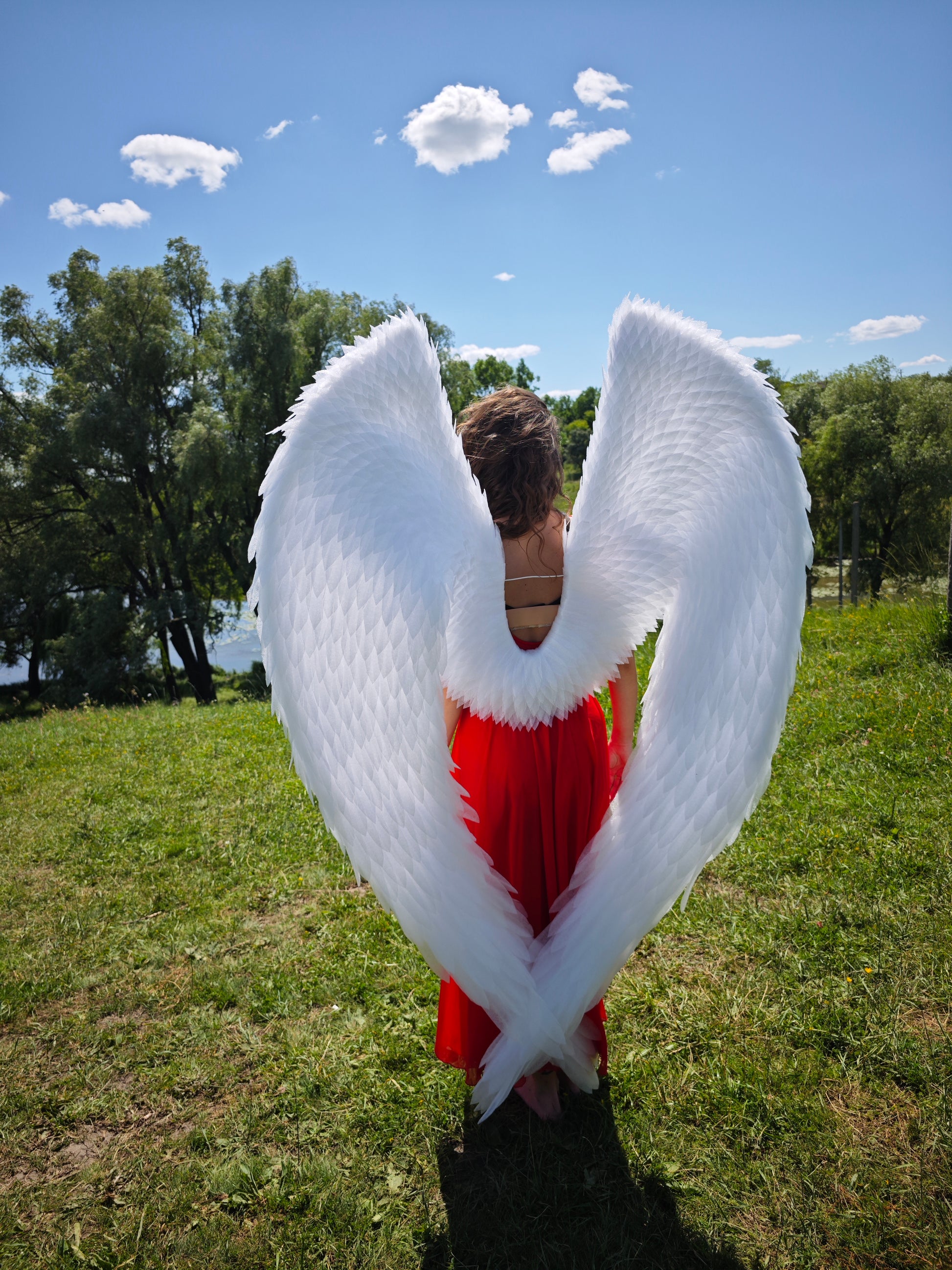 Person wearing large white angel wings outdoors on a sunny day with trees and grass.